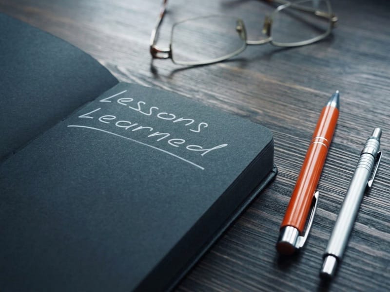 Notebook open to a dark page with Lessons Learned written on it, beside two pens and glasses