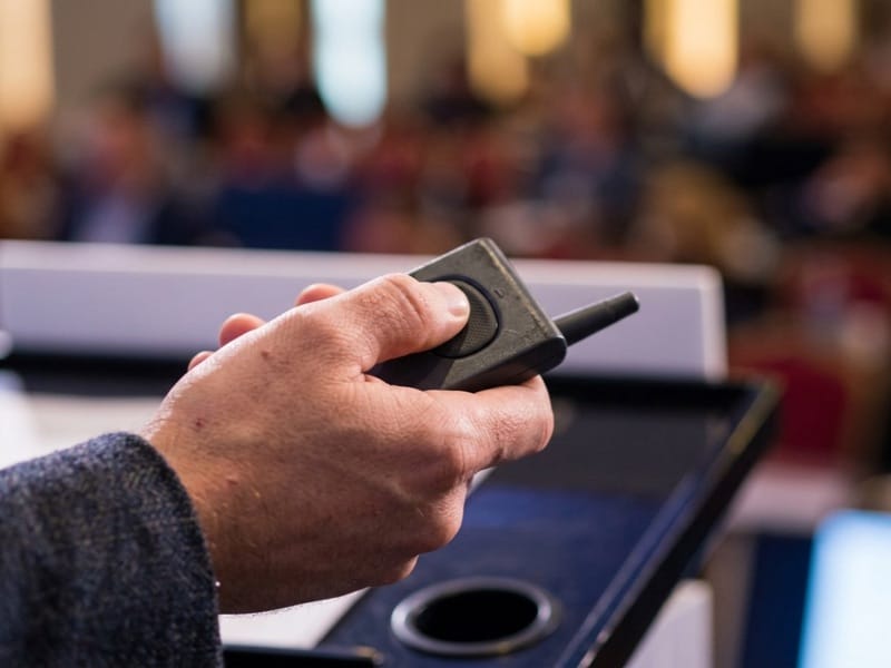 Hand holding a presenter clicker at a lectern in front of a blurred conference audience