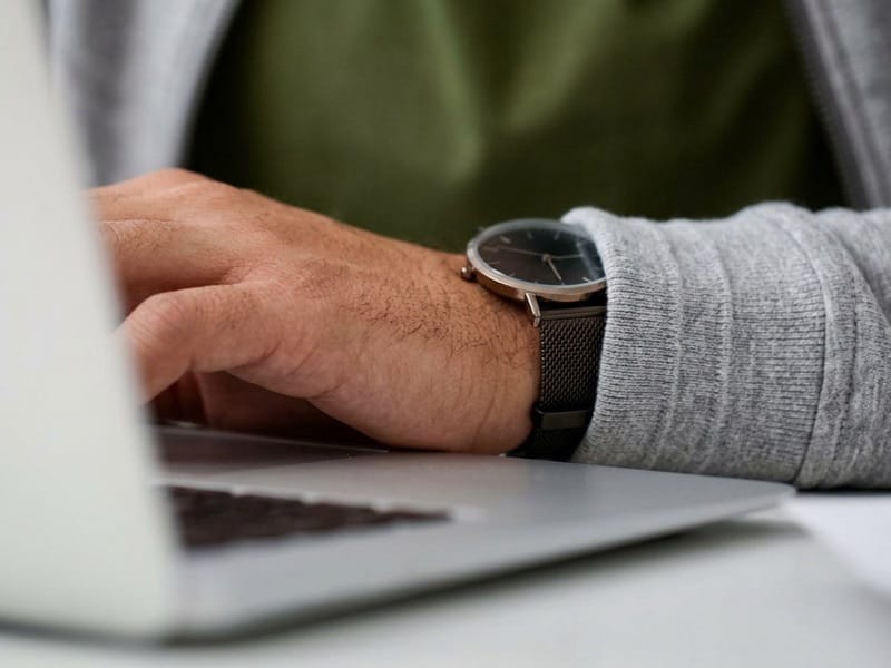 Man wearing a wristwatch and grey cardigan typing on a laptop