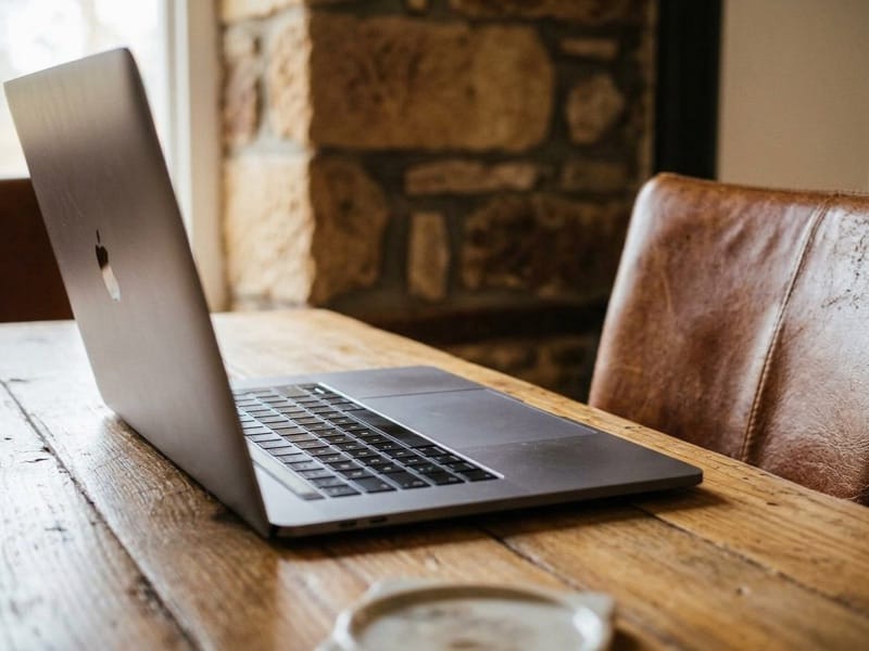 Open laptop on a rustic wooden table with a leather chair and stone wall behind