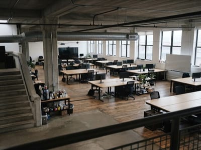 An empty open-plan office interior with wooden desks, computer monitors, office chairs, concrete pillars, large windows, and a concrete staircase.