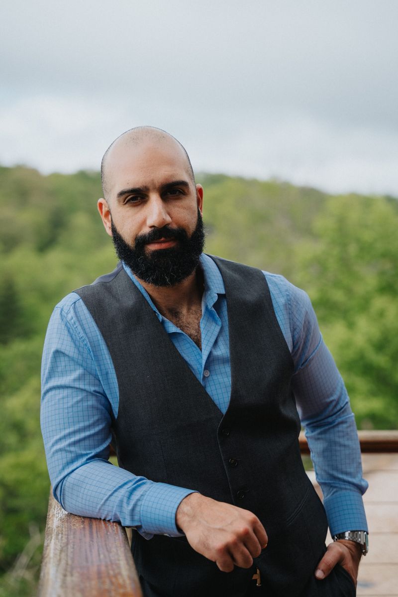 Portrait of Ayman Al-Abdullah in a vest leaning on a wooden railing with green hills behind