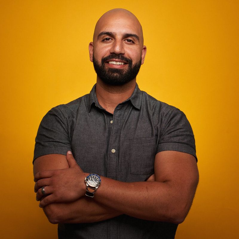 Studio headshot of Ayman Al-Abdullah, smiling with his arms crossed against a yellow backdrop