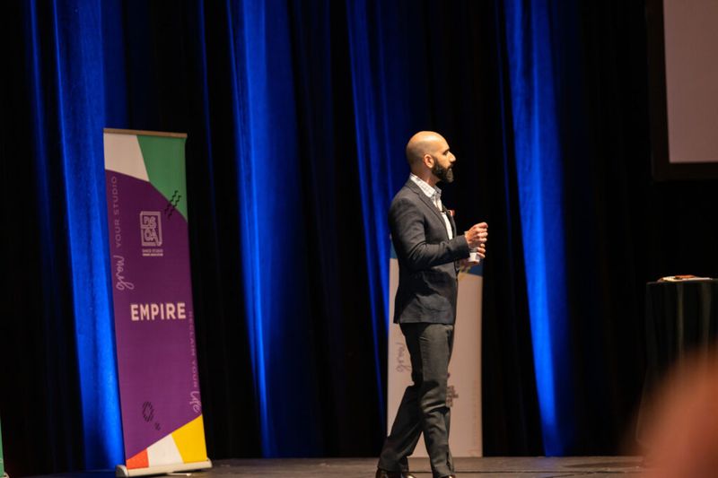 Ayman Al-Abdullah speaking on stage beside a Dance Studio Owners Association Empire banner