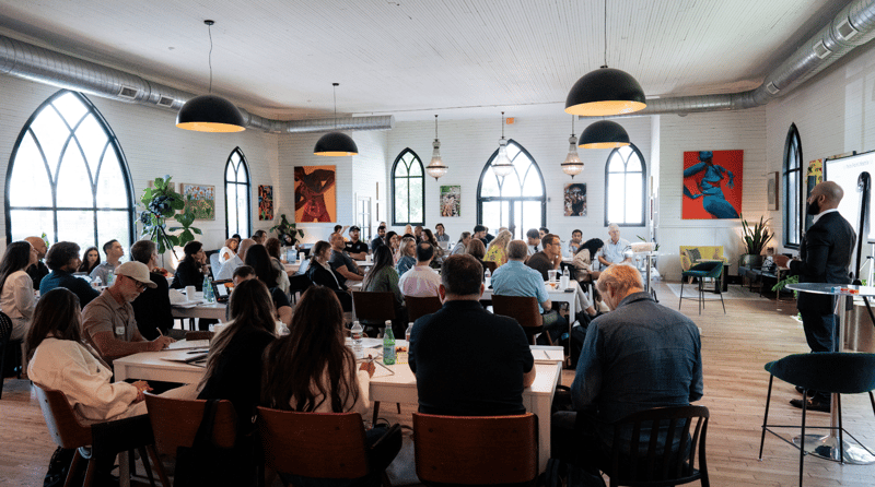 A workshop in progress in a sunlit loft with arched windows, attendees seated at tables and a presenter at the front