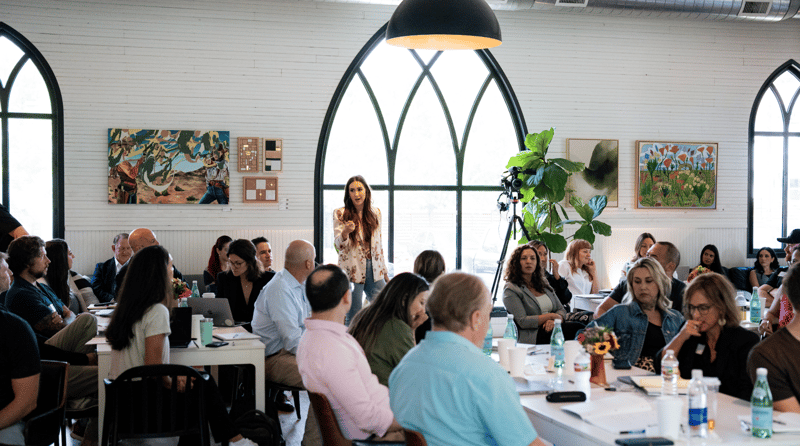 A speaker addressing workshop attendees seated at tables in a sunlit room with arched windows
